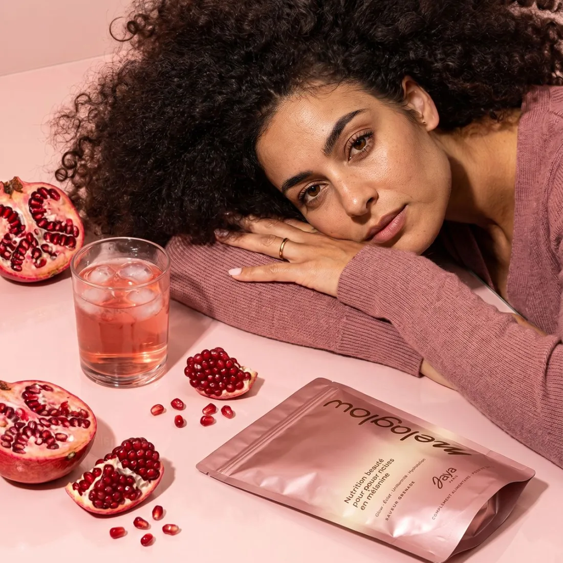 A woman resting her head on her arms beside a glass, a MelaGlow pouch and open pomegranates