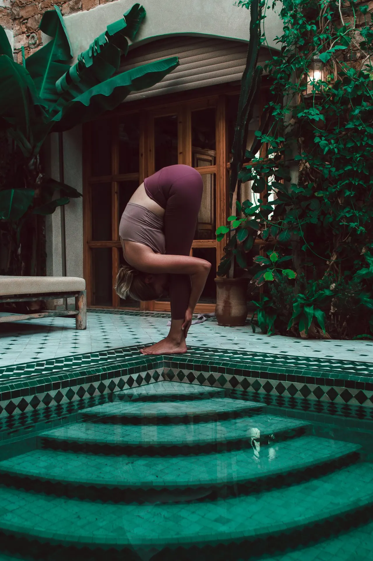 A woman in a yoga forward fold beside a tiled pool